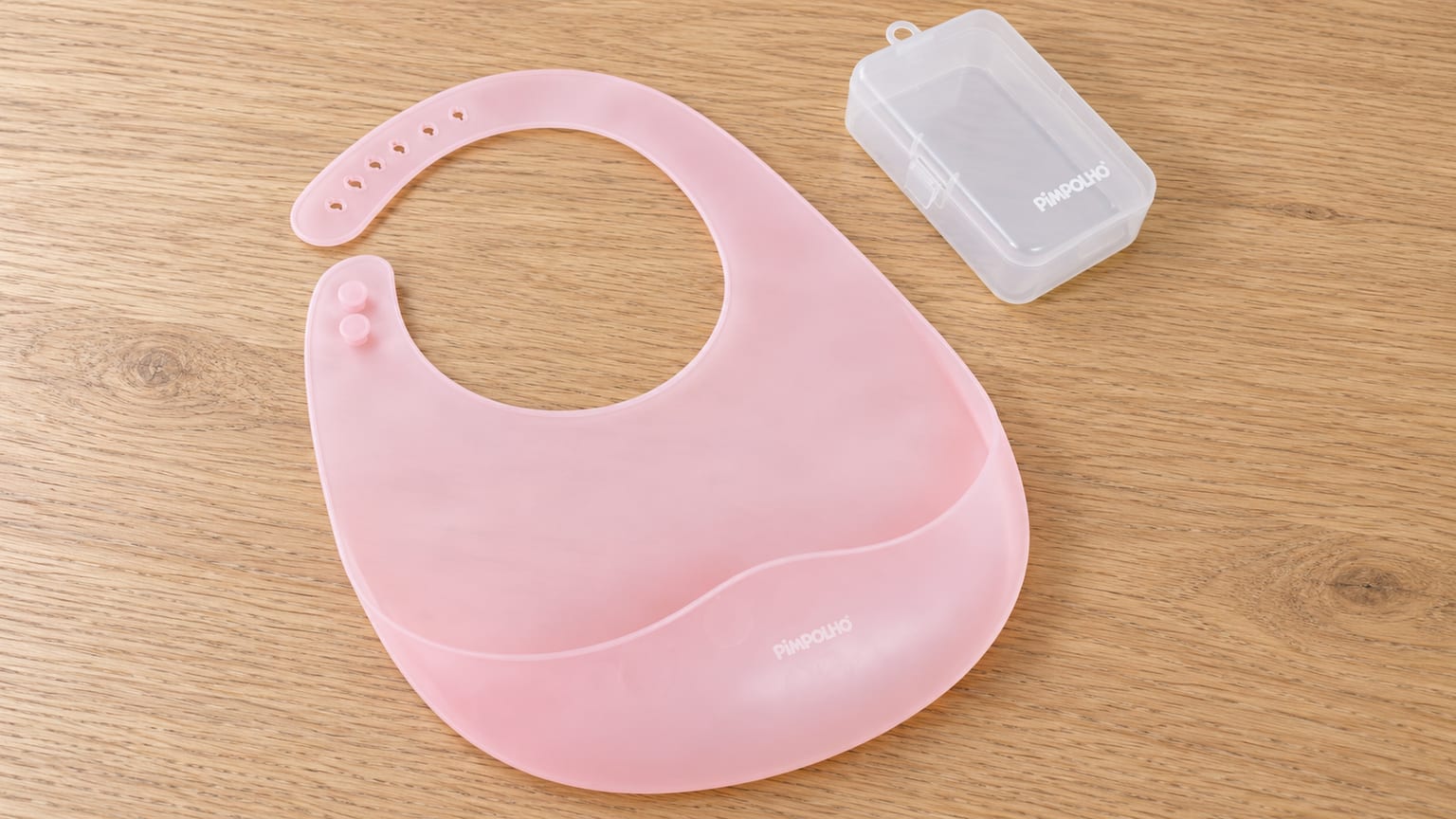 Babador Pimpolho rosa com bolso coletor, ao lado do estojo translúcido, sobre uma mesa de madeira clara.