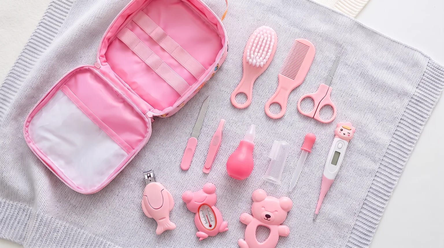 Bolsa rosa aberta e acessórios de cuidados do bebê sobre uma manta cinza, com escova, pente, tesoura, termômetro digital e termômetro de banho em formato de sapo.