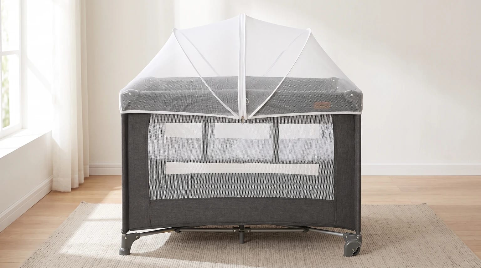 Berço portátil cinza com mosquiteiro branco em formato de tenda, montado sobre um tapete em um quarto bem iluminado.