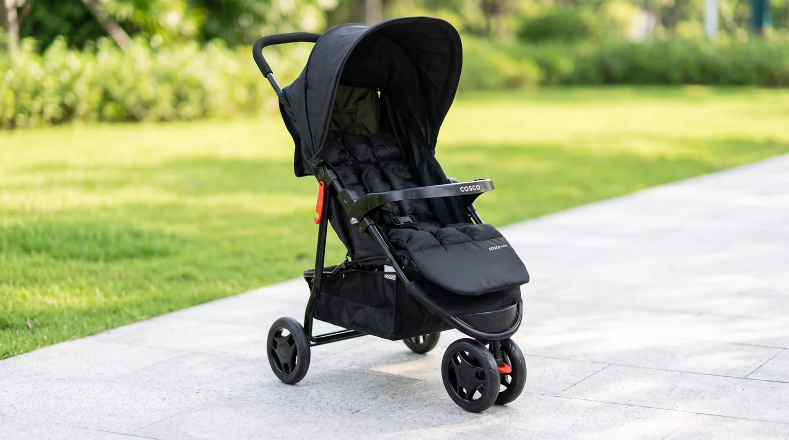 Carrinho de bebê Cosco todo preto, com assento acolchoado, parado em um caminho de parque ao lado da grama.