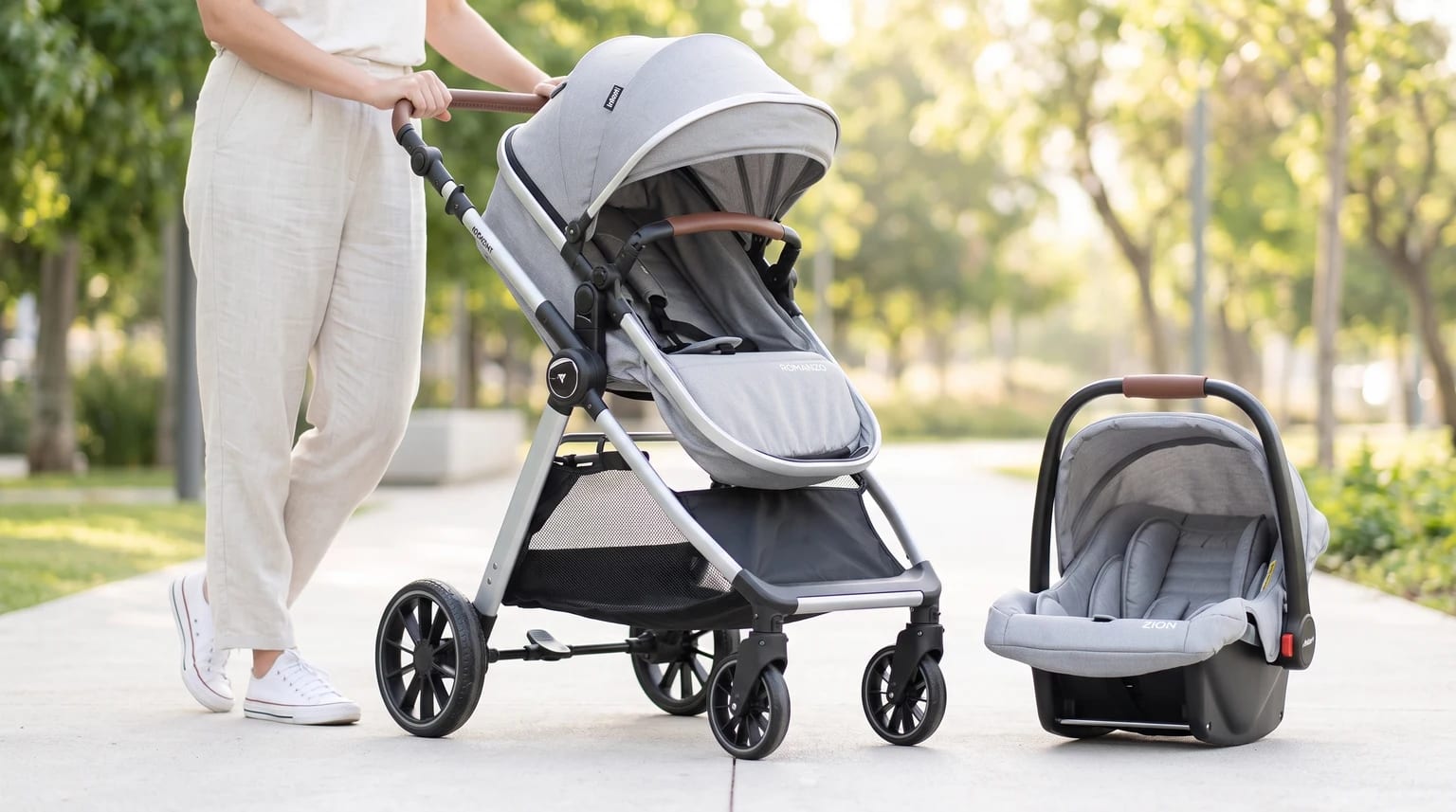 Uma mulher ao lado de um carrinho de bebê Infanti Romanzo Duo cinza e do bebê conforto correspondente em um caminho de parque.