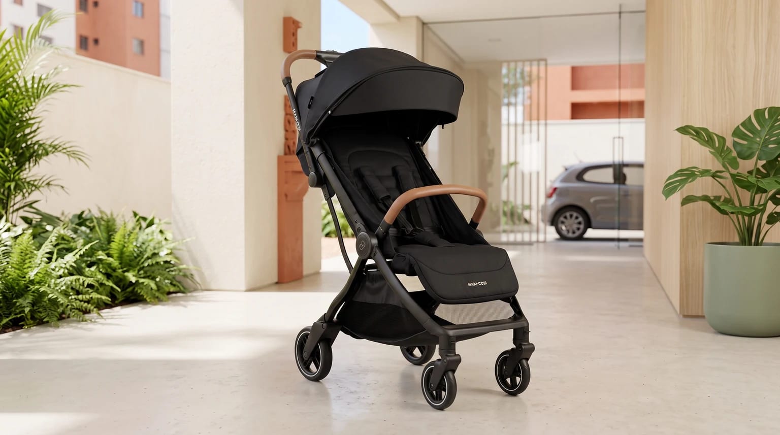 Carrinho de bebê Maxi-Cosi Eva³ preto, com alça e barra de segurança marrons, em uma entrada de condomínio iluminada.
