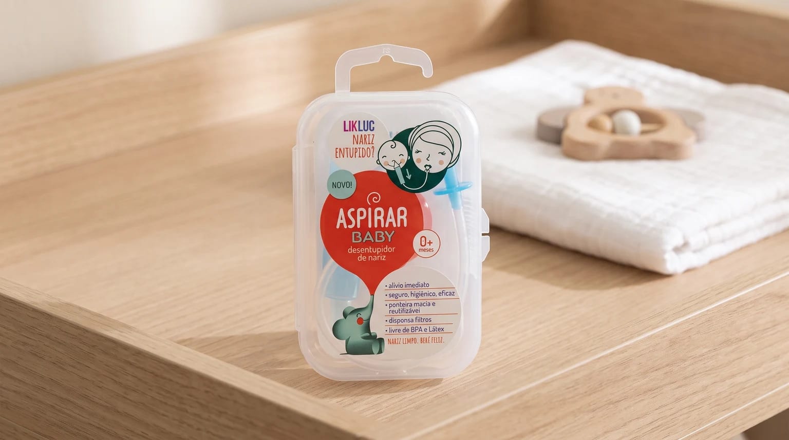 Estojo do aspirador nasal Likluc AspirarBaby sobre um trocador de madeira, ao lado de um pano branco e um chocalho.