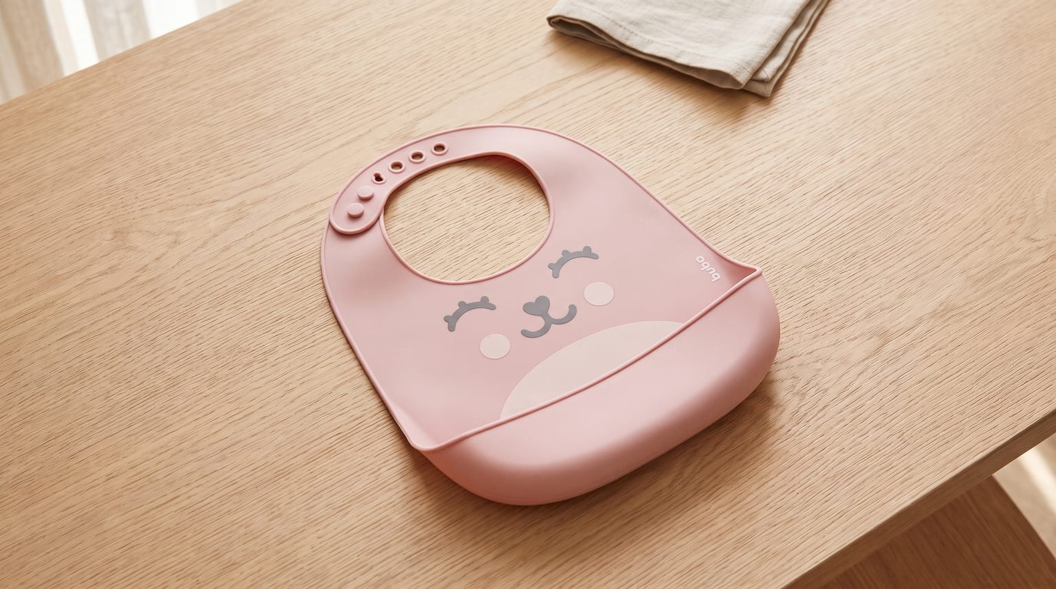 Babador de silicone rosa com um rostinho de animal estampado, sobre uma mesa de madeira clara.
