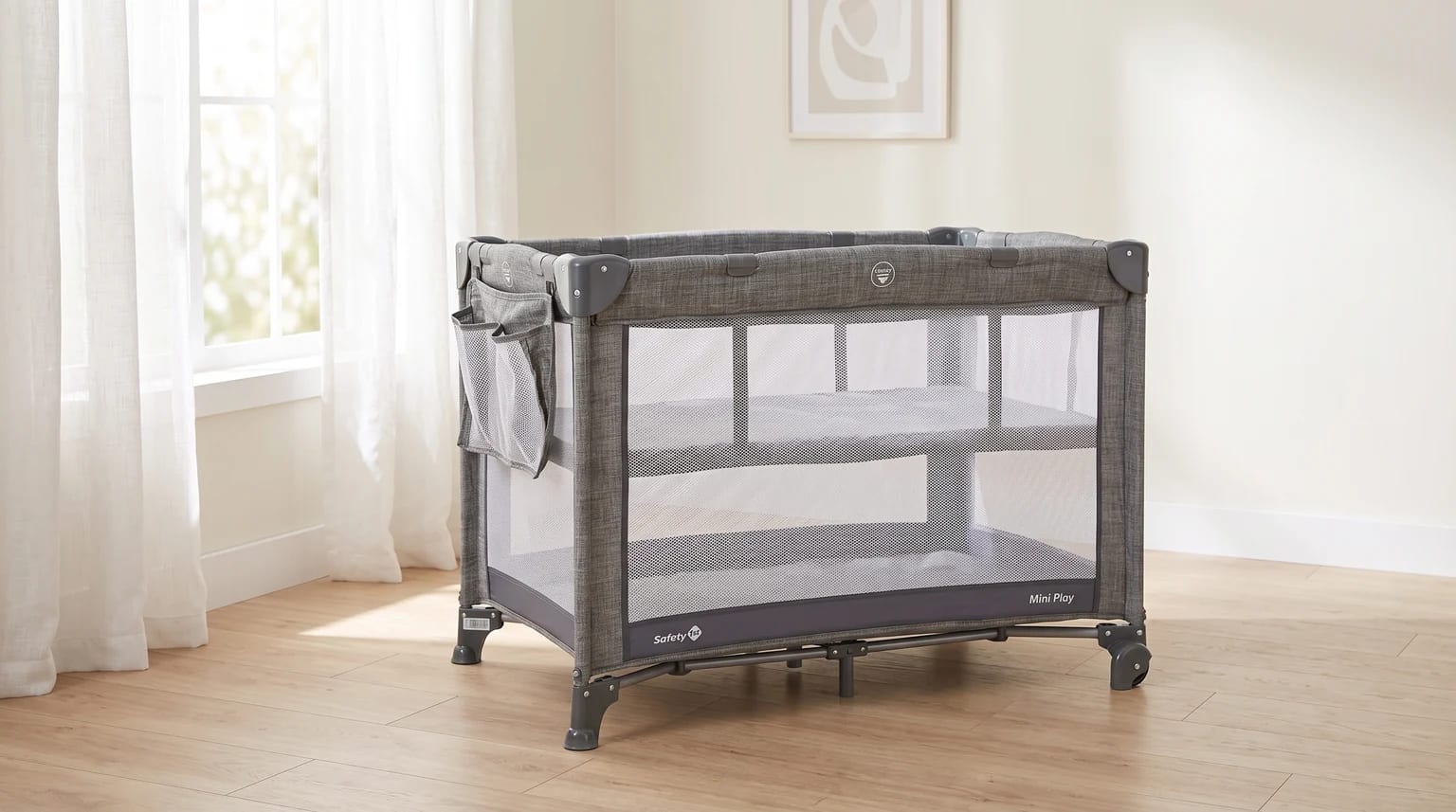 Berço portátil cinza da Safety 1st montado em um quarto com piso de madeira, ao lado de uma janela com cortinas brancas.