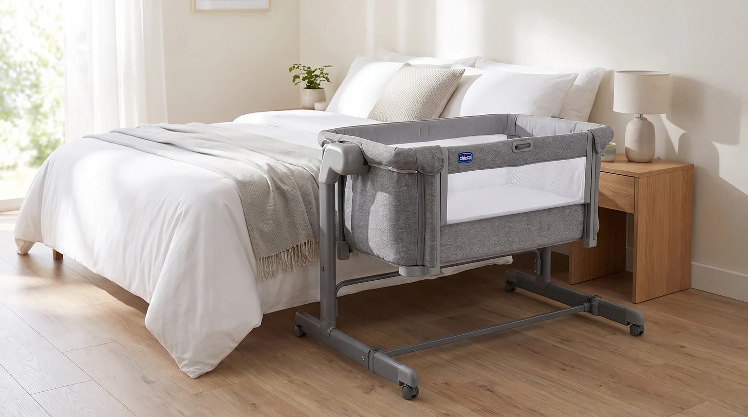 Berço Chicco Next2Me Magic Evo cinza com rodas, ao lado de uma cama de casal.