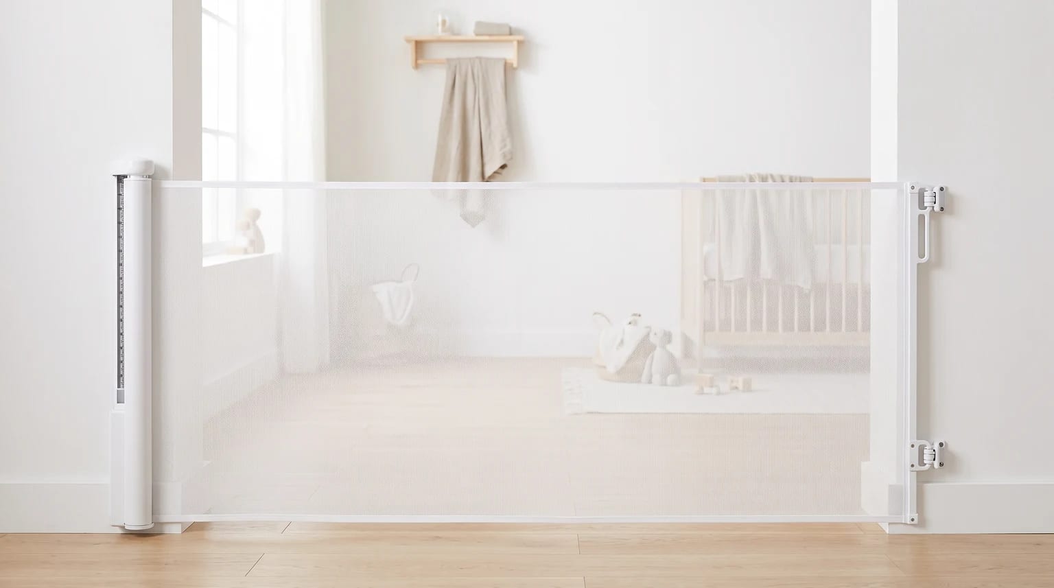 Um portão de segurança retrátil de malha branca instalado na passagem para um quarto de bebê, com um berço e brinquedos ao fundo.