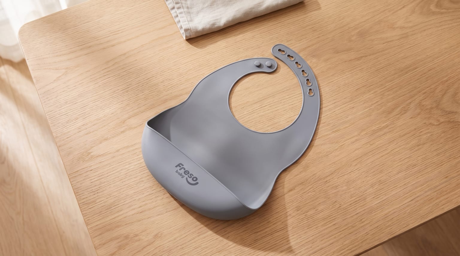 Babador de silicone cinza com bolso coletor e fecho ajustável, deitado sobre uma mesa de madeira clara.