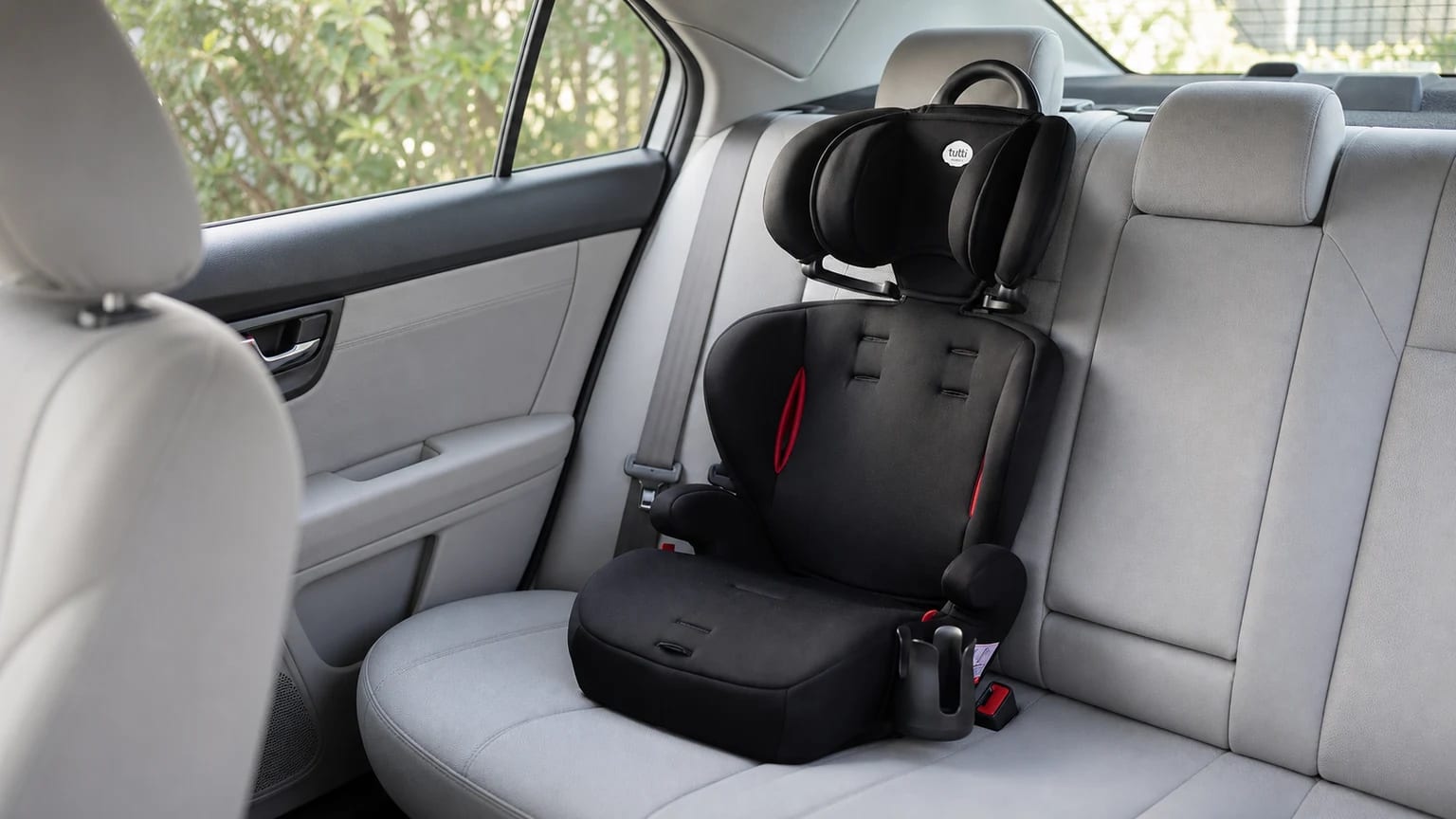 Cadeirinha Tutti Baby Versati preta com detalhes vermelhos e porta-copo, instalada no banco traseiro de um carro com interior cinza-claro.