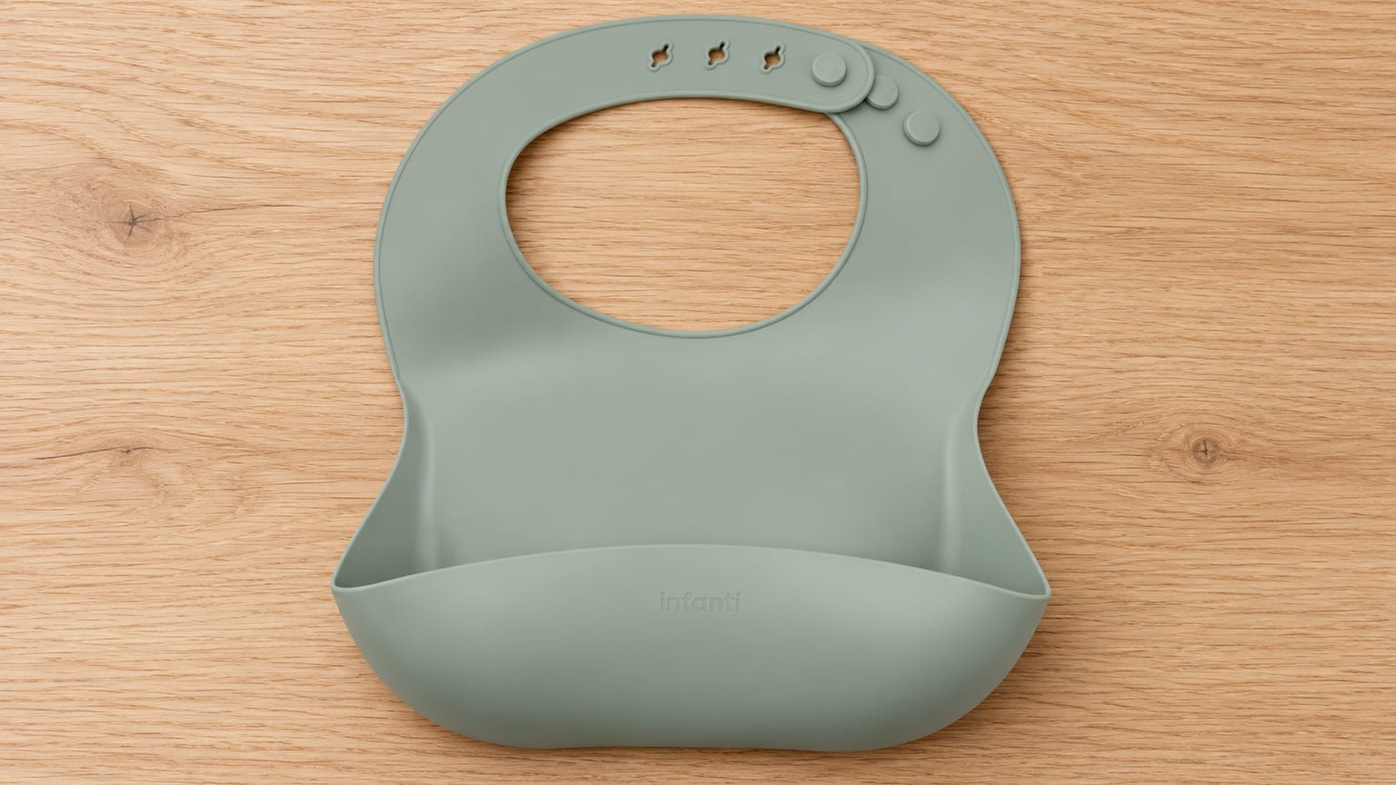 Babador Infanti Piccolo verde com bolso coletor e fecho ajustável, sobre uma mesa de madeira clara.