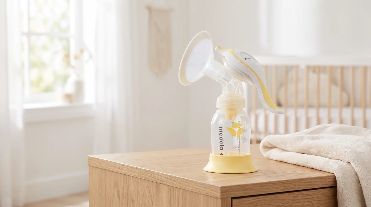 Bombinha manual branca e amarela da Medela sobre uma cômoda de madeira em um quarto de bebê, com um berço ao fundo.