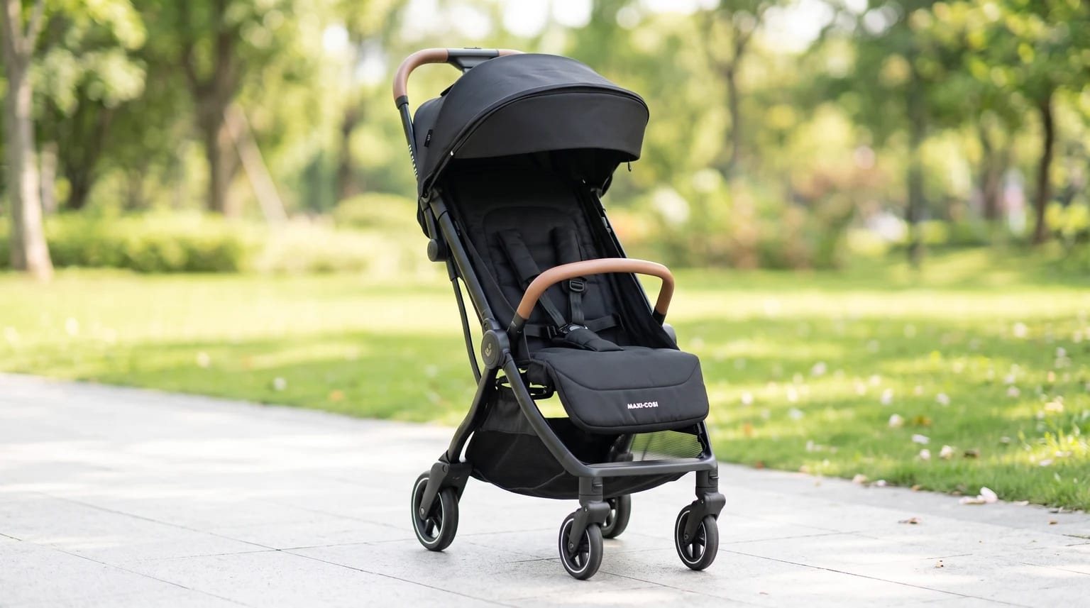Carrinho de bebê preto com alça e barra frontal marrons em um caminho de parque com árvores ao fundo.