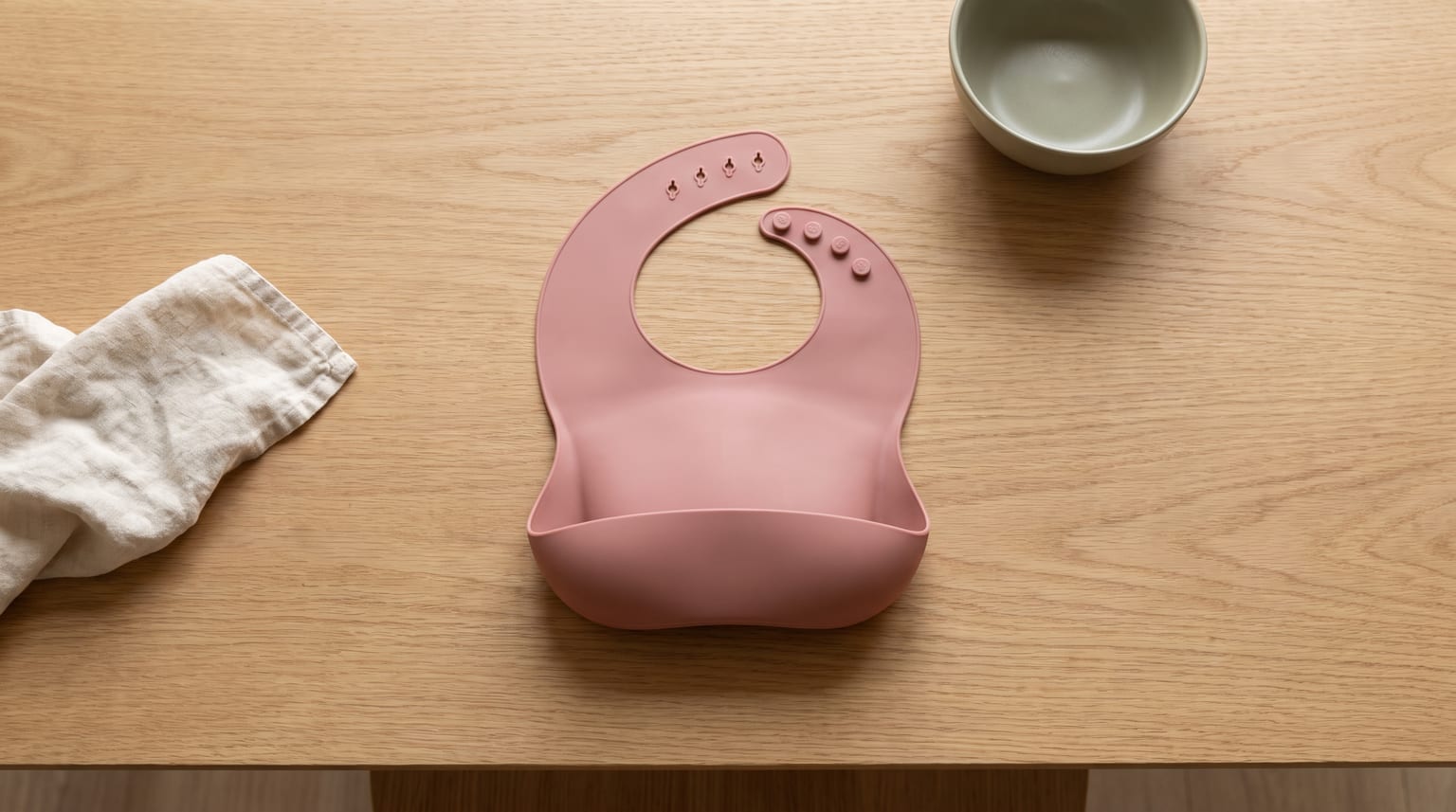 Um babador de silicone rosa sobre uma mesa de madeira, ao lado de uma pequena tigela e de um guardanapo de pano.